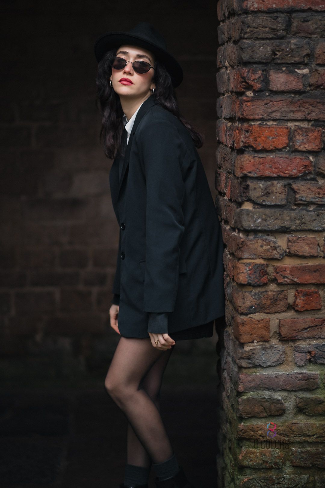 Stylish figure in black blazer, hat and sunglasses poses dramatically against brick wall in moody outdoor portrait.