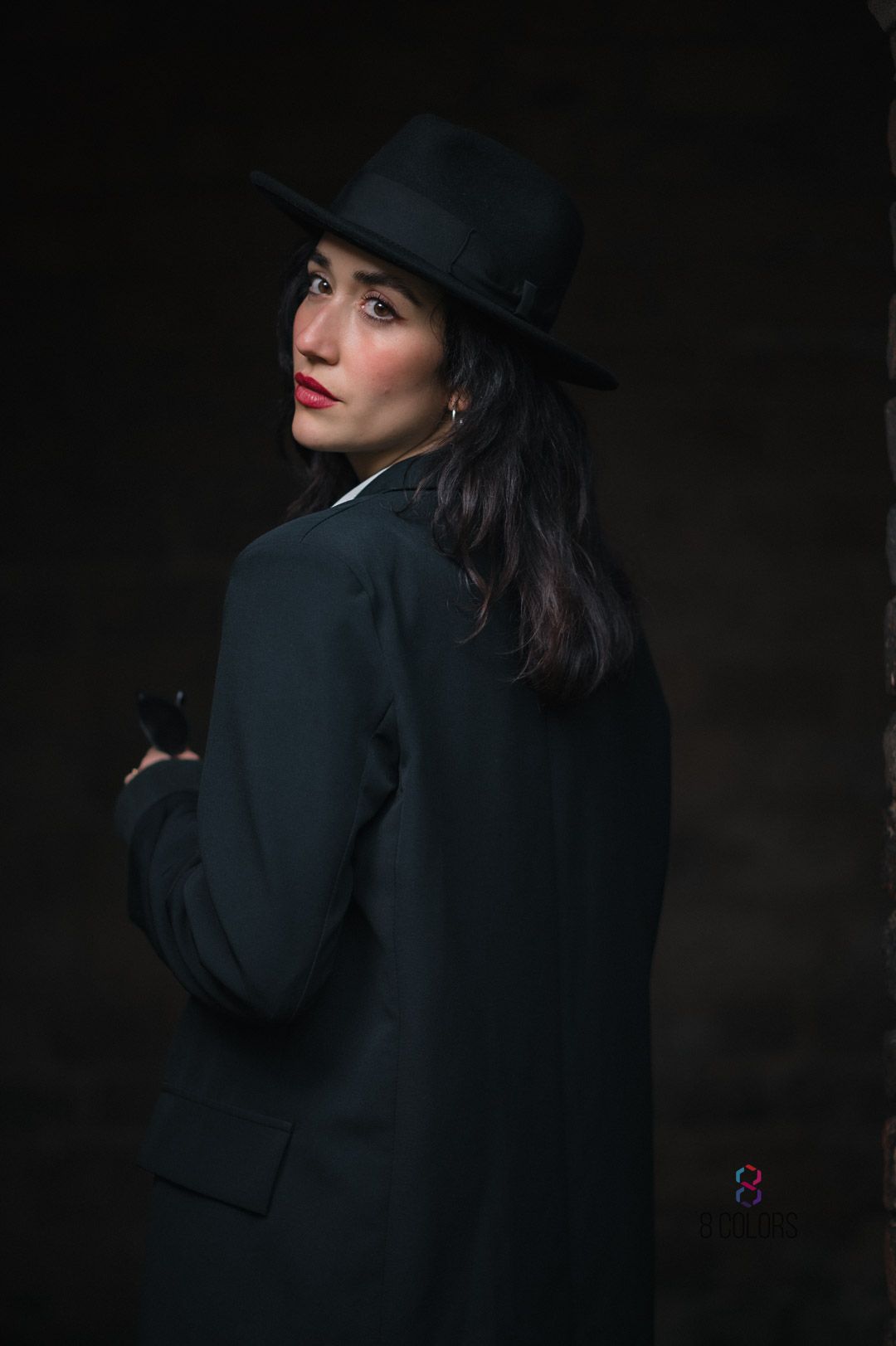 Dramatic portrait in dark lighting features a figure in black coat and fedora hat looking over their shoulder against dark background.