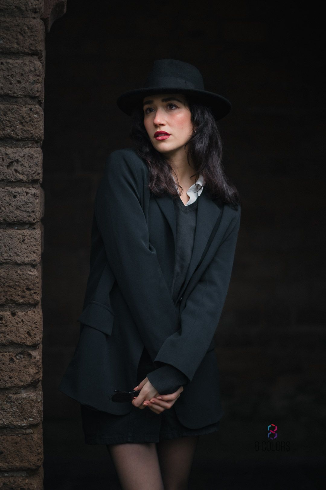 Moody portrait of figure in dark blazer and fedora hat against brick wall with dramatic lighting and shadows.