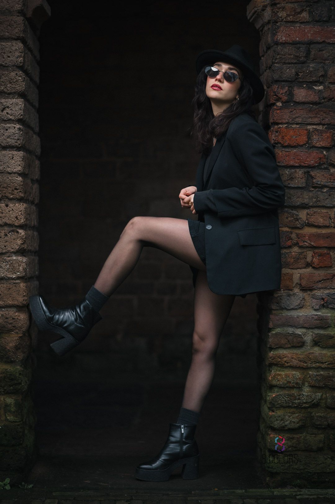 Fashion portrait of a person in dark oversized blazer, black hat and combat boots posing against brick wall background.
