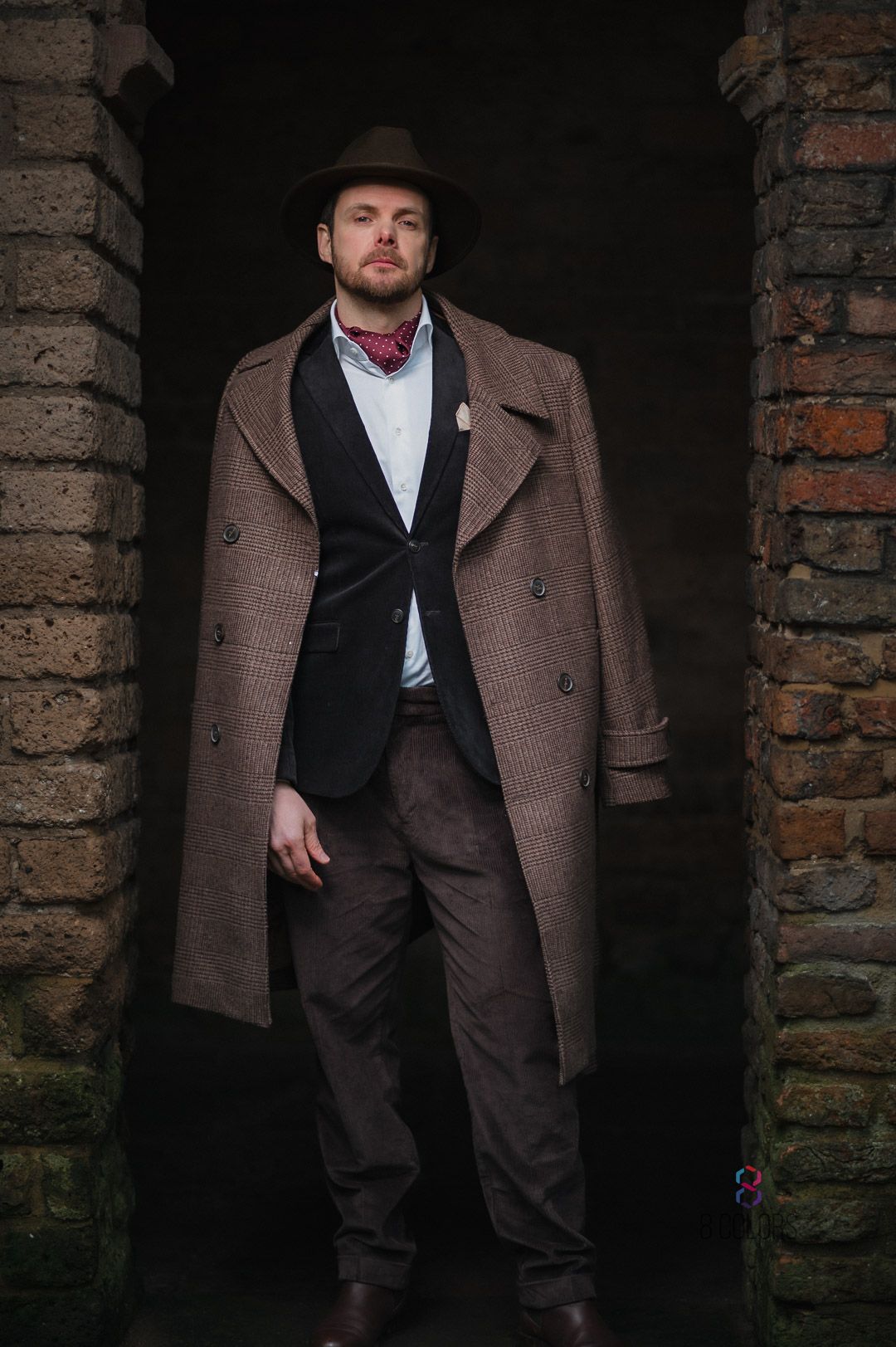 Stylishly dressed figure in brown overcoat and hat poses confidently between brick archway in moody lighting.