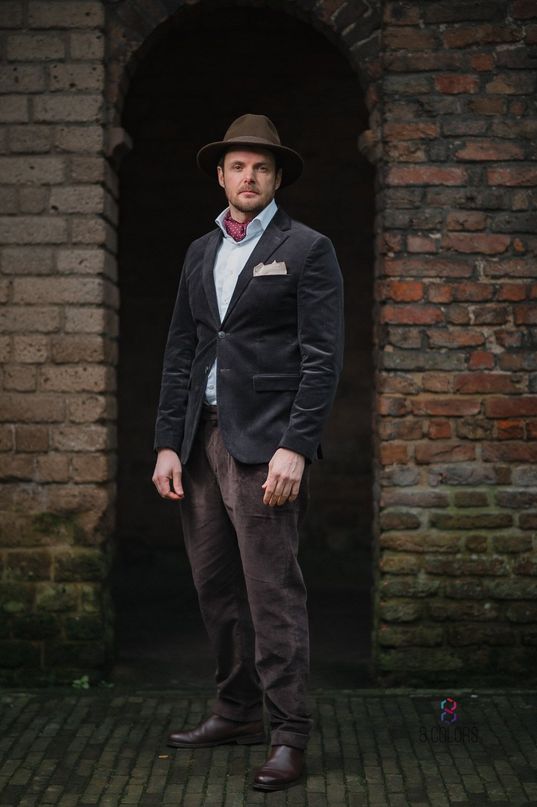Stylish velvet blazer and fedora outfit photographed in front of rustic brick archway in urban setting.