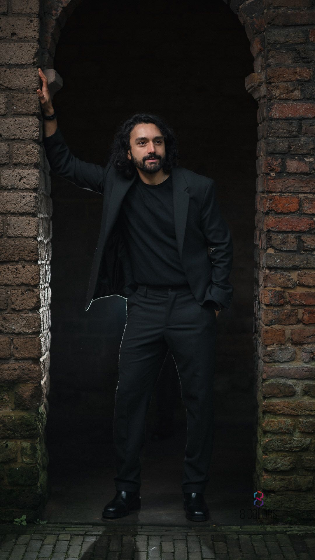 A person wearing all black attire poses against a brick archway in dramatic lighting.