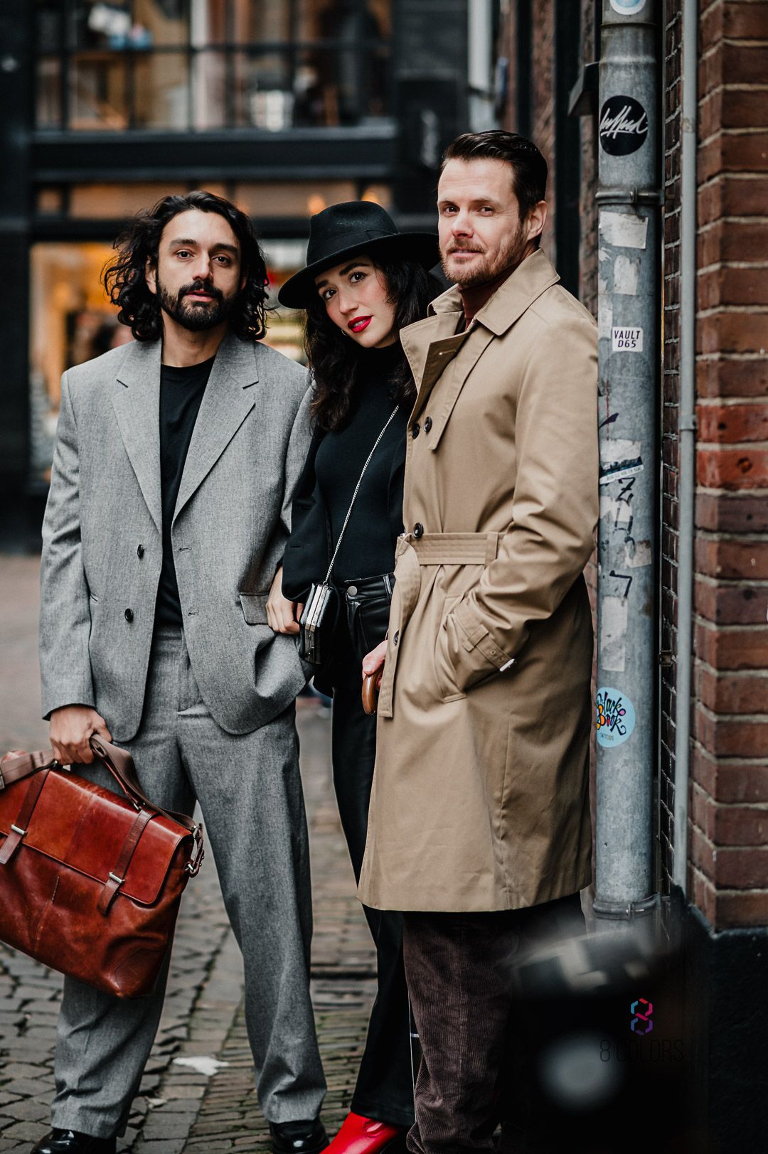 Three people in trendy outfits posing against brick wall on cobblestone street in city.