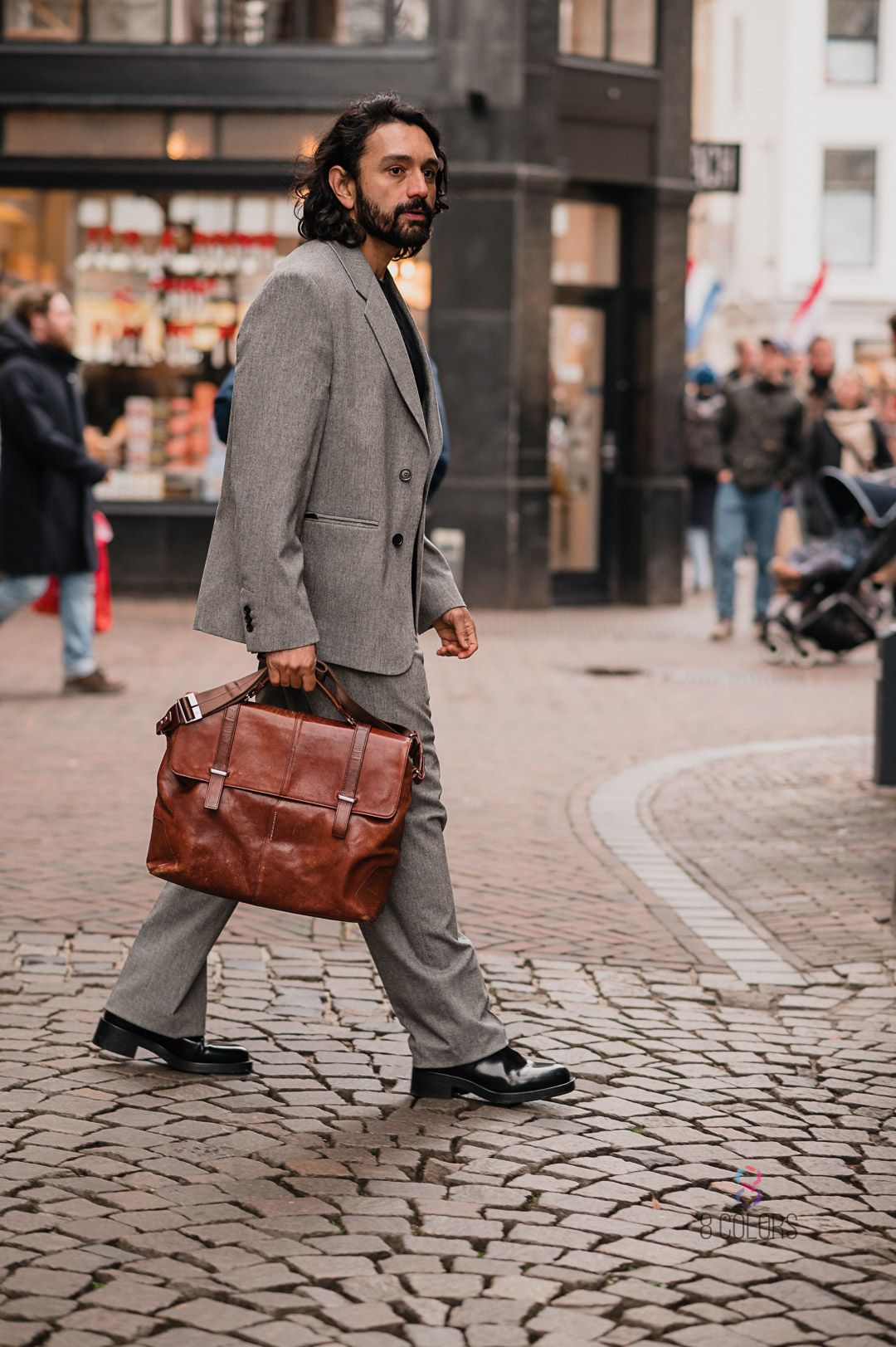 Stylishly dressed person in grey suit walking on cobblestone street carrying brown leather briefcase in urban setting.