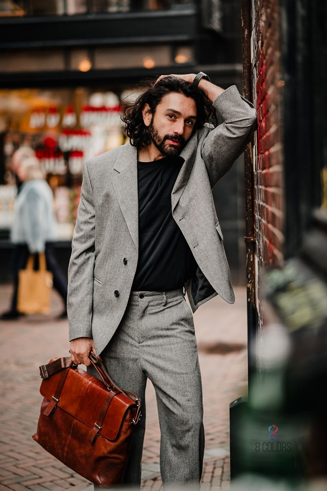 Fashionable person in gray suit with leather briefcase posing against brick wall on urban street.
