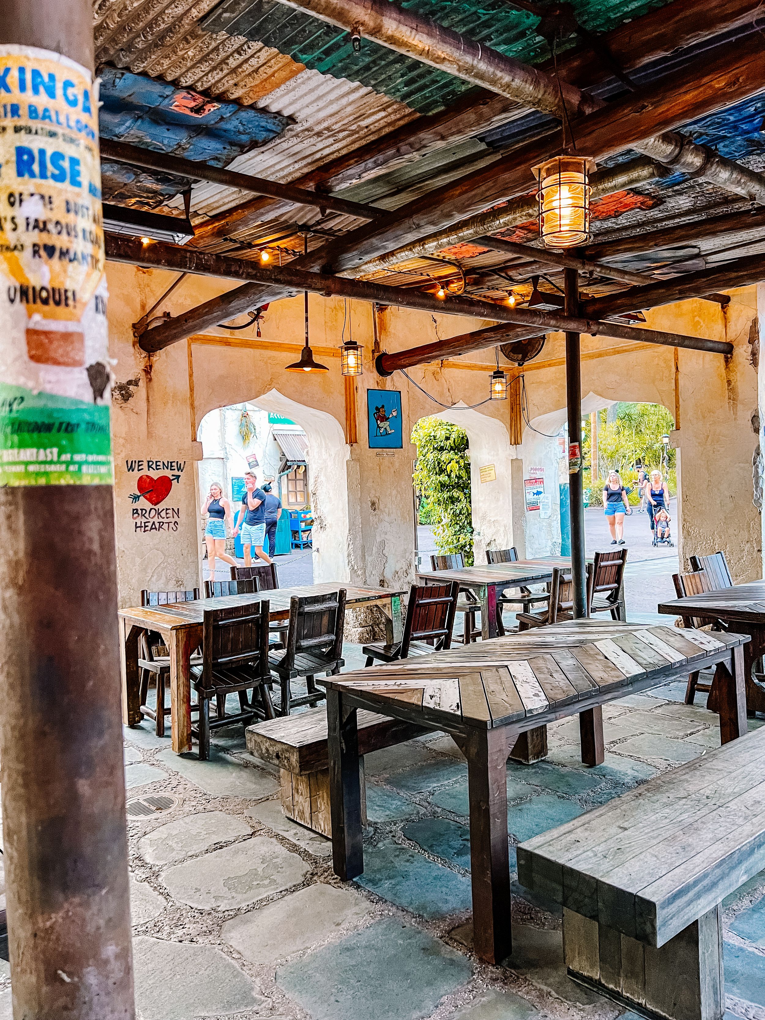 Rustic outdoor dining area with wooden benches and hanging lanterns under a weathered covered patio in a tropical setting.