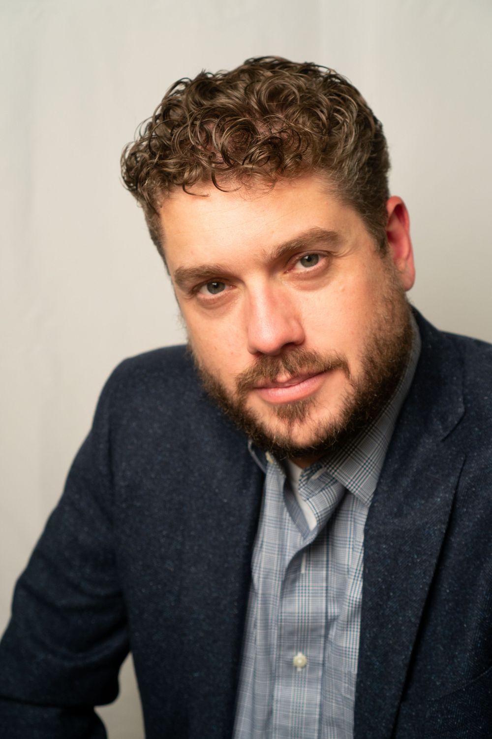 Professional headshot of person in navy blazer and checkered shirt against light background.