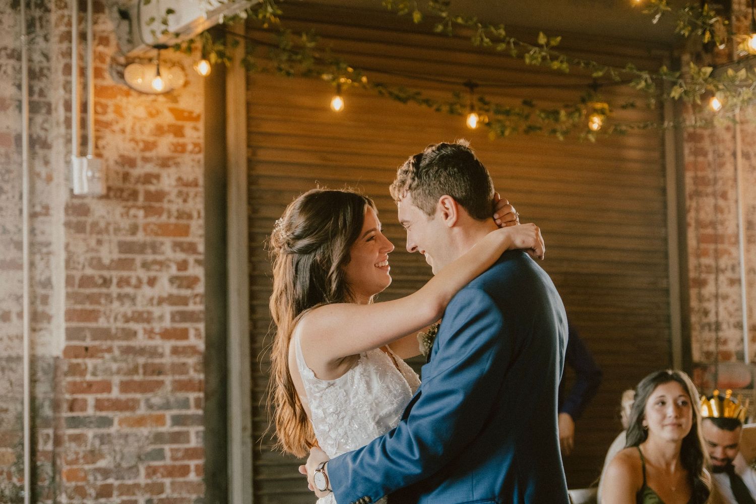 Couple dances in romantic embrace at wedding reception with exposed brick walls and string lights.