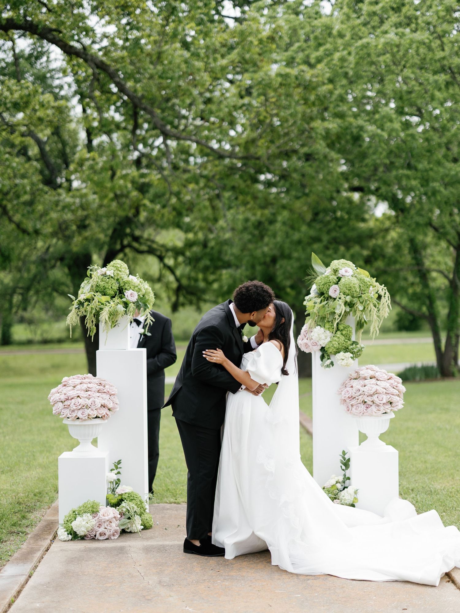 Romantic wedding ceremony outdoors with white floral arrangements and pedestals framing the couple and officiant.