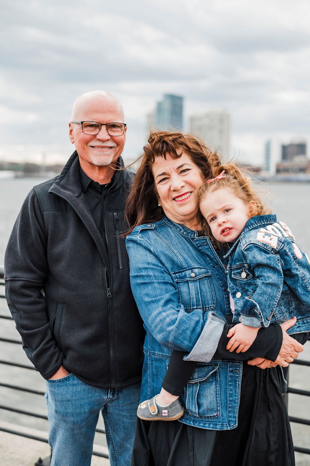 Three generations pose together on a pier with a city skyline visible in the background on an overcast day.
