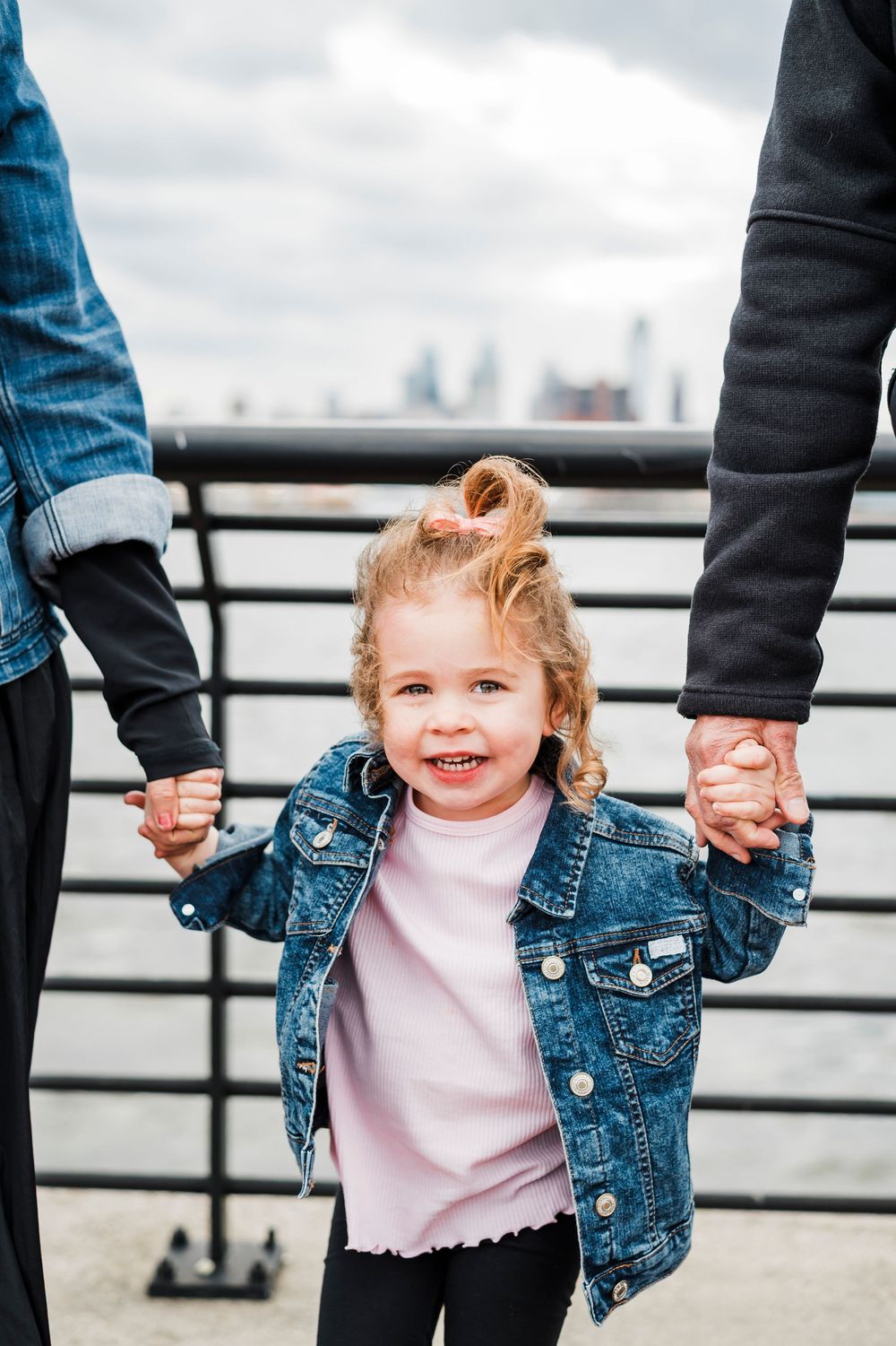 Small child wearing denim jacket holds hands with adults against urban skyline backdrop on waterfront.