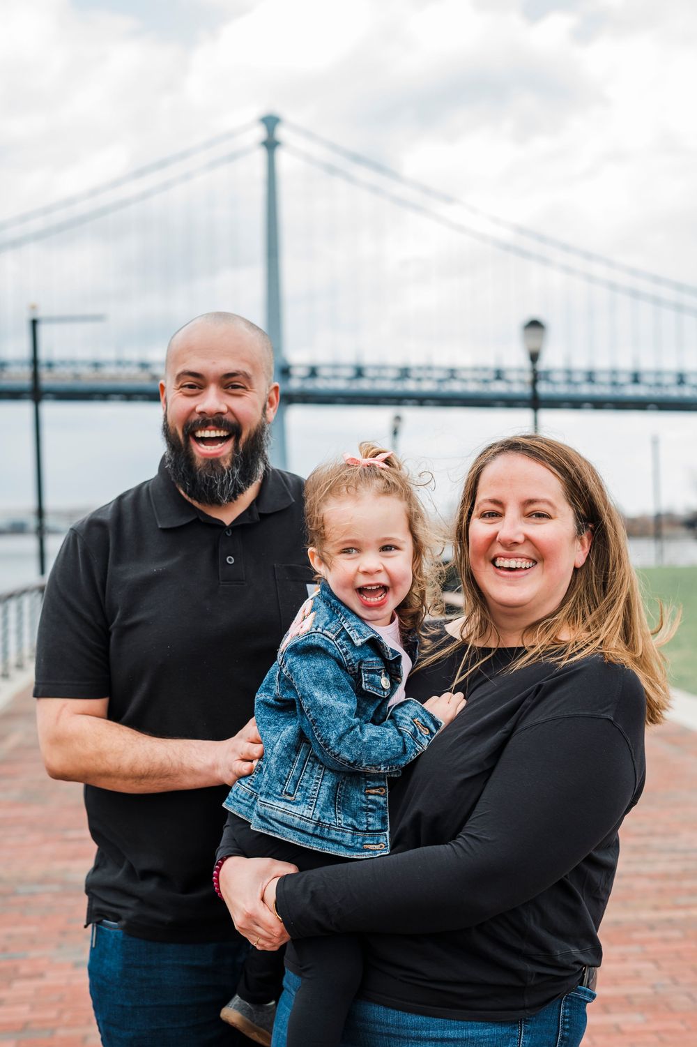 A happy family smiles together while standing near a bridge on a brick walkway on an overcast day.