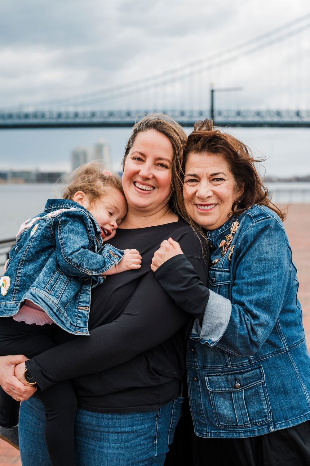 Three people share a warm embrace outside on a cloudy day with a bridge visible in the background.