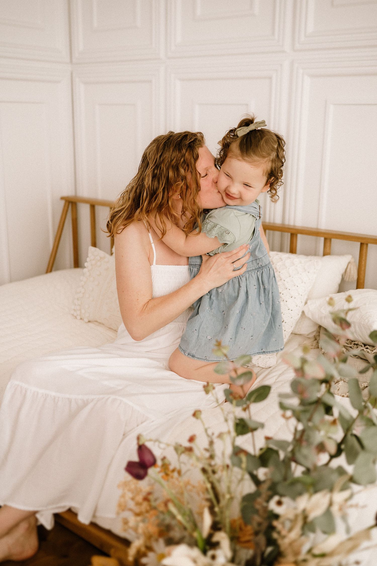 Sweet tender moment between parent and child on white bedding with dried floral arrangement displayed beside them.