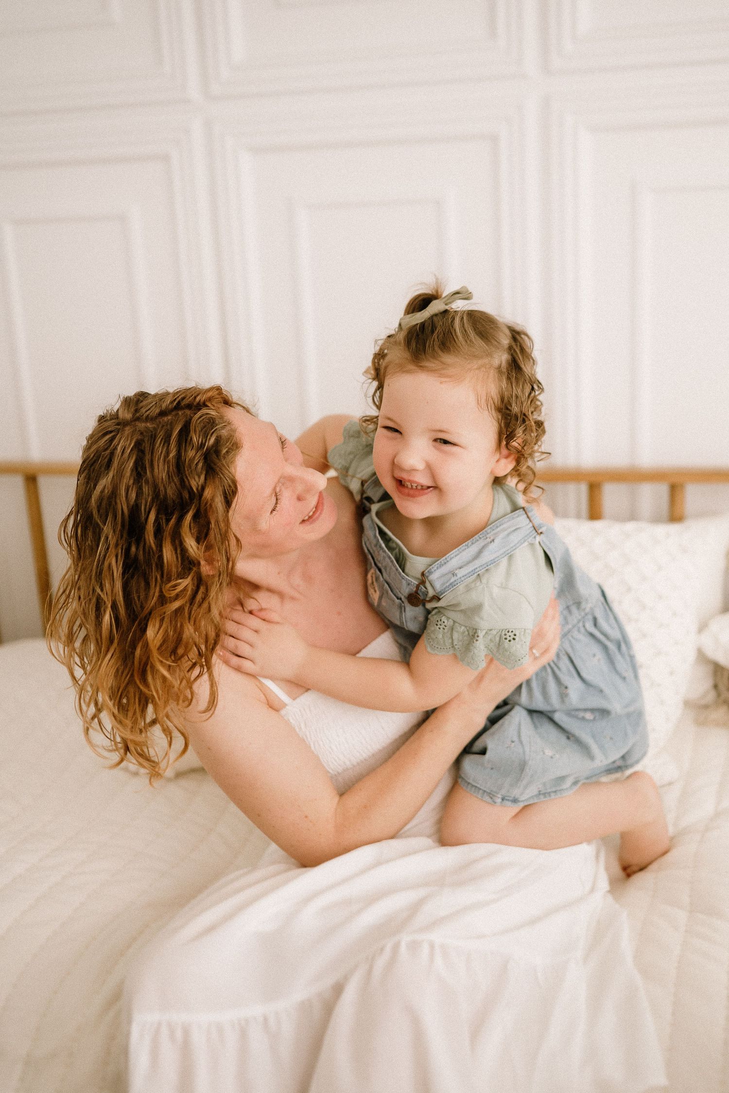 A loving moment captured between mother and child laughing together on white bedding in a bright room.