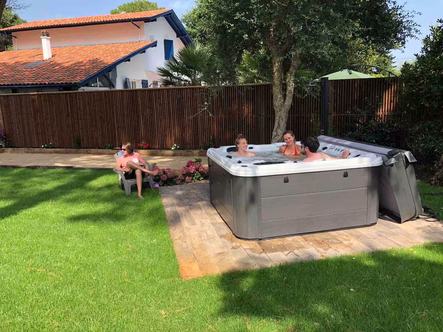People relax in a backyard hot tub and on lounge chairs near a wooden fence on a sunny summer day.
