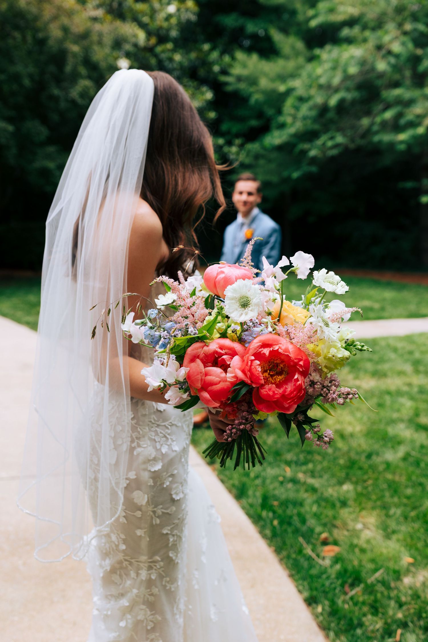 A wedding bouquet with vibrant coral peonies and white flowers against a lush green outdoor backdrop.