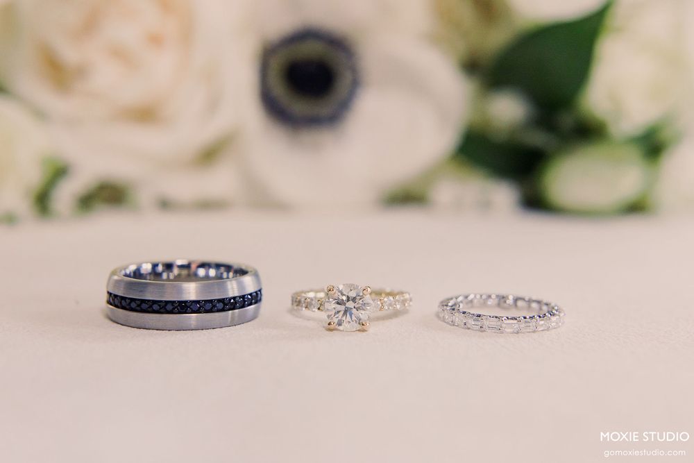 Wedding rings and bands displayed on a soft white surface with blurred floral decorations in the background.