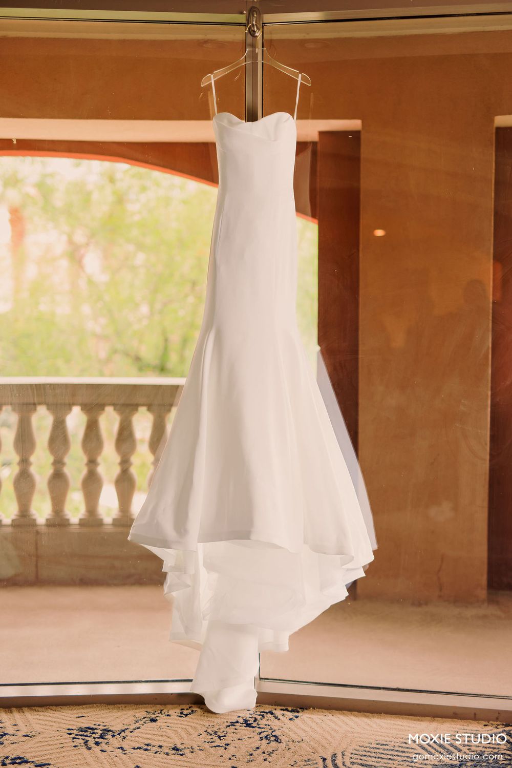 A white wedding dress hangs elegantly against a warm-toned wall with decorative balustrades visible in the background.