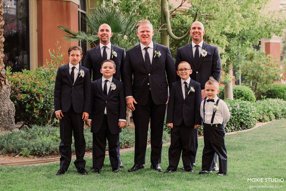 Wedding party in formal black attire poses together on green lawn in front of brick building.