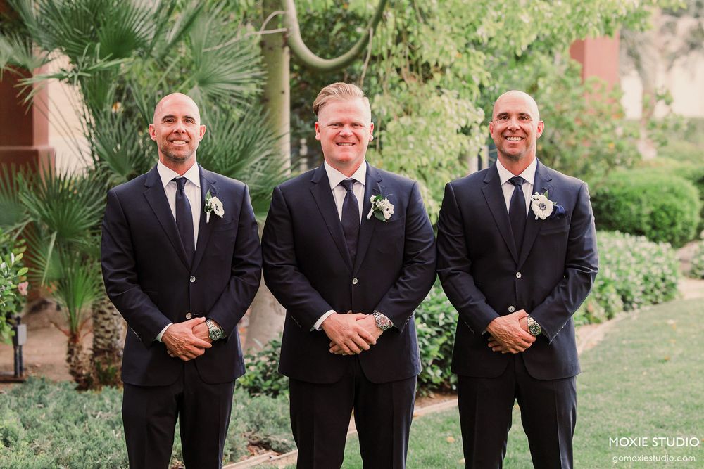 Three groomsmen in black tuxedos and matching boutonnieres pose together outdoors in a garden setting.