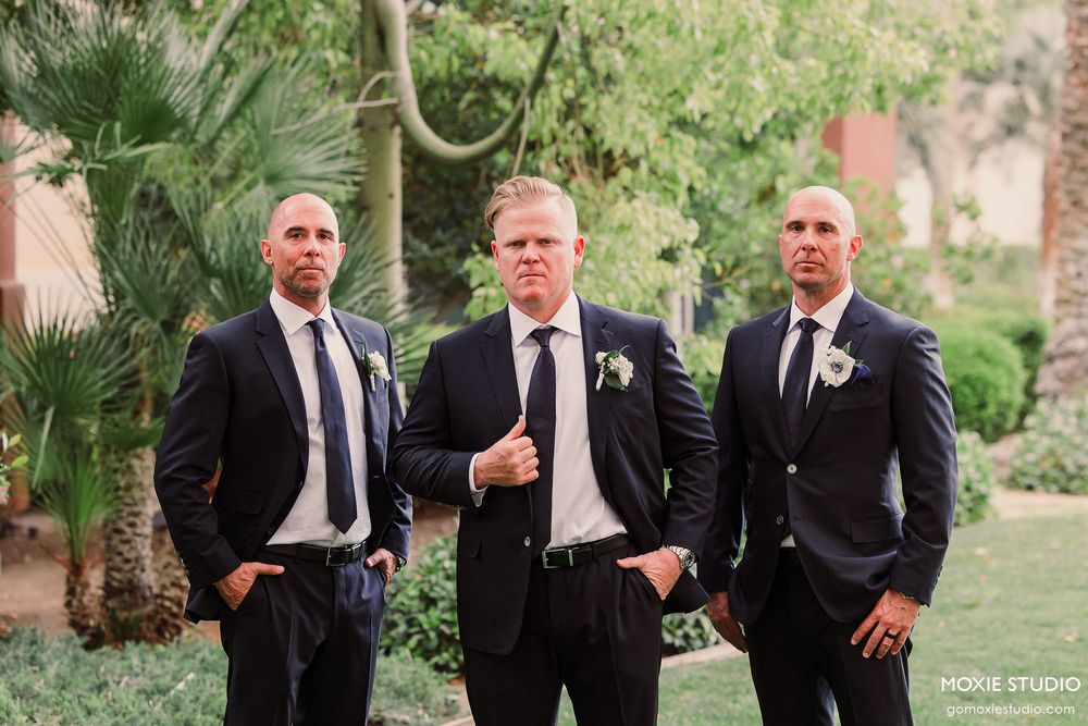 Groomsmen in black suits pose together in a lush tropical garden setting during a wedding celebration.