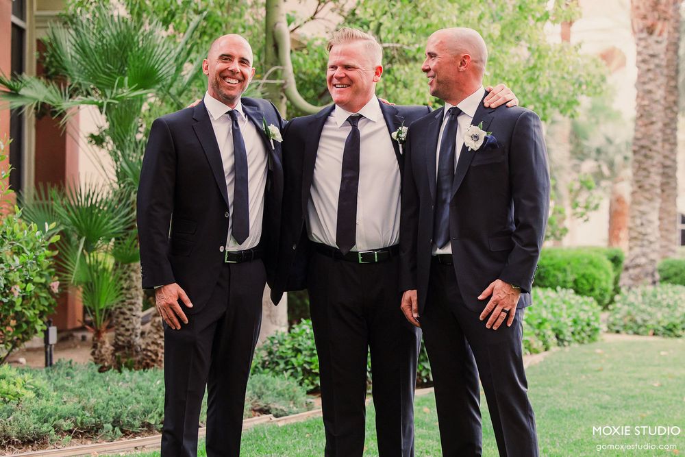 Groomsmen in black suits share a lighthearted moment outdoors surrounded by lush greenery and palm trees.