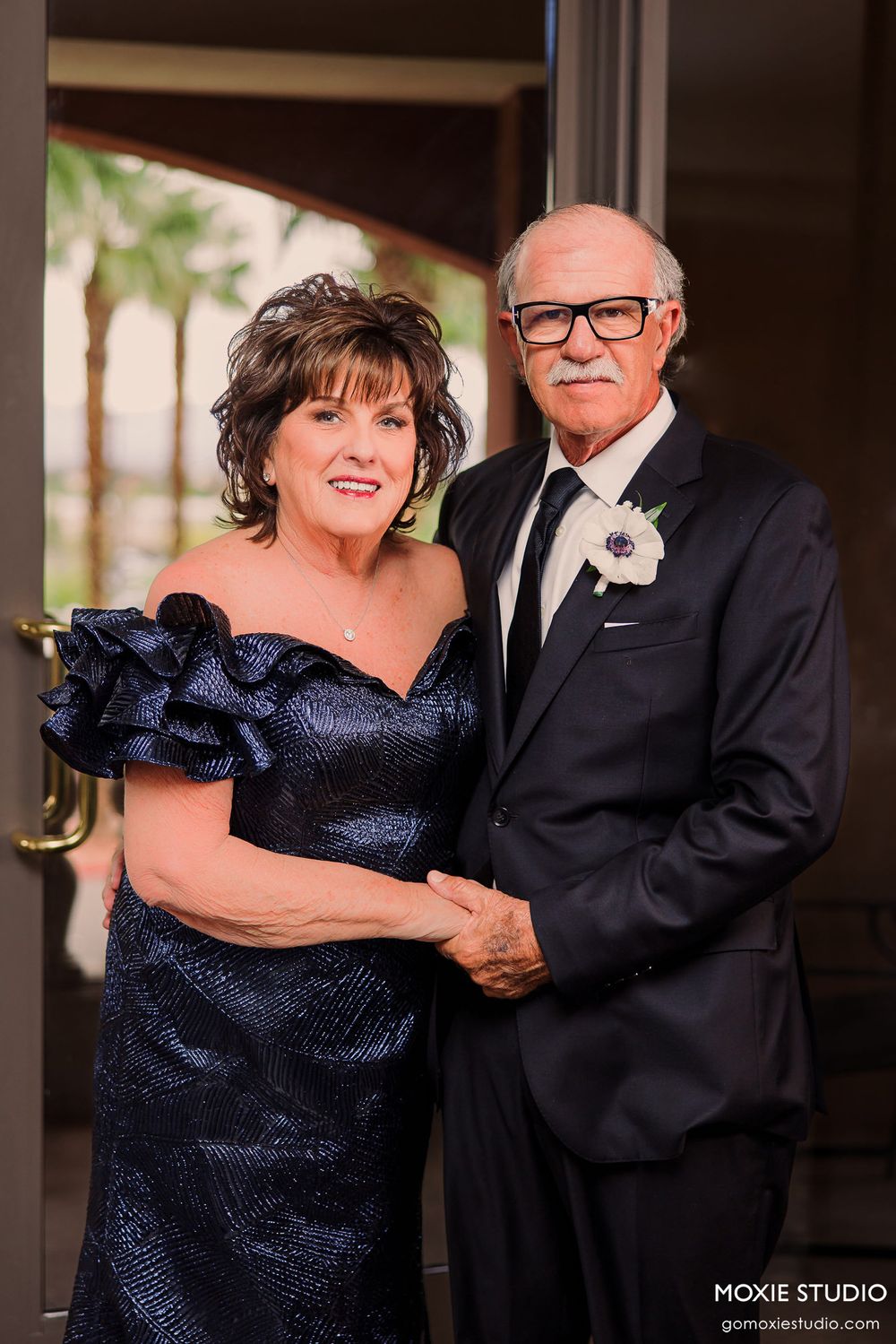 Couple in formal attire pose together at an event, with woman in navy dress and man in black tuxedo with glasses.