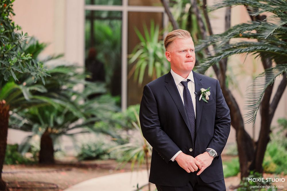 Men in formal black suits stand in an outdoor garden setting with tropical plants and palm trees.