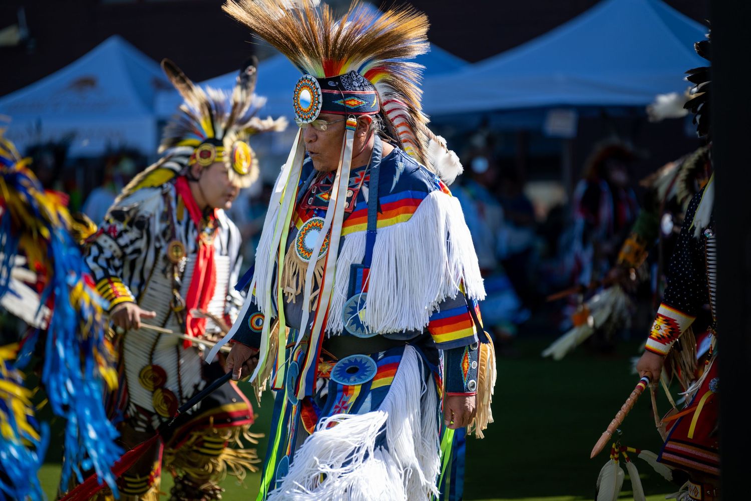 Ceremonial dancers in feathered headdresses and colorful regalia perform at an outdoor gathering.
