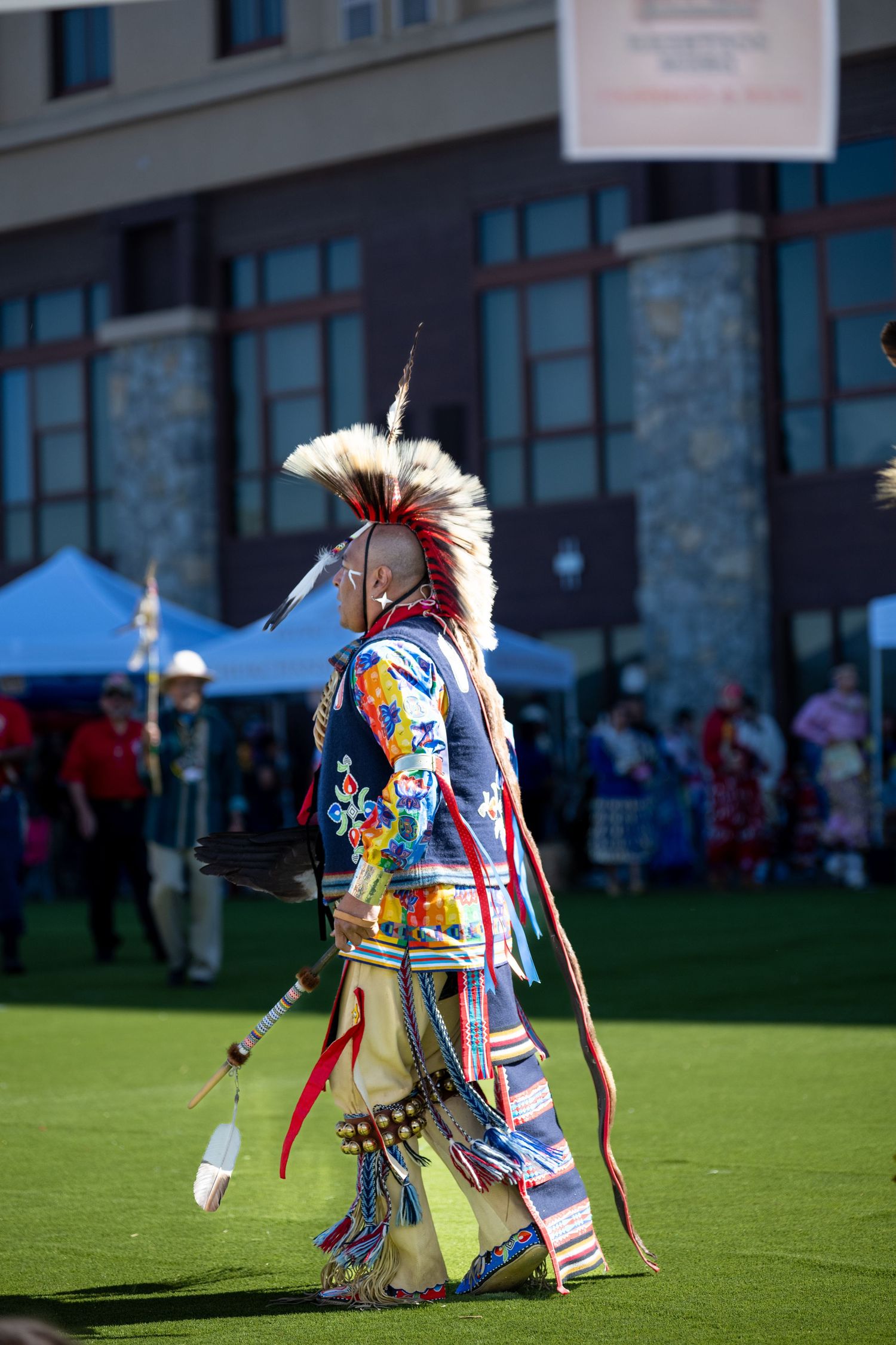 Dancers in traditional regalia perform on a grassy field with buildings in the background.