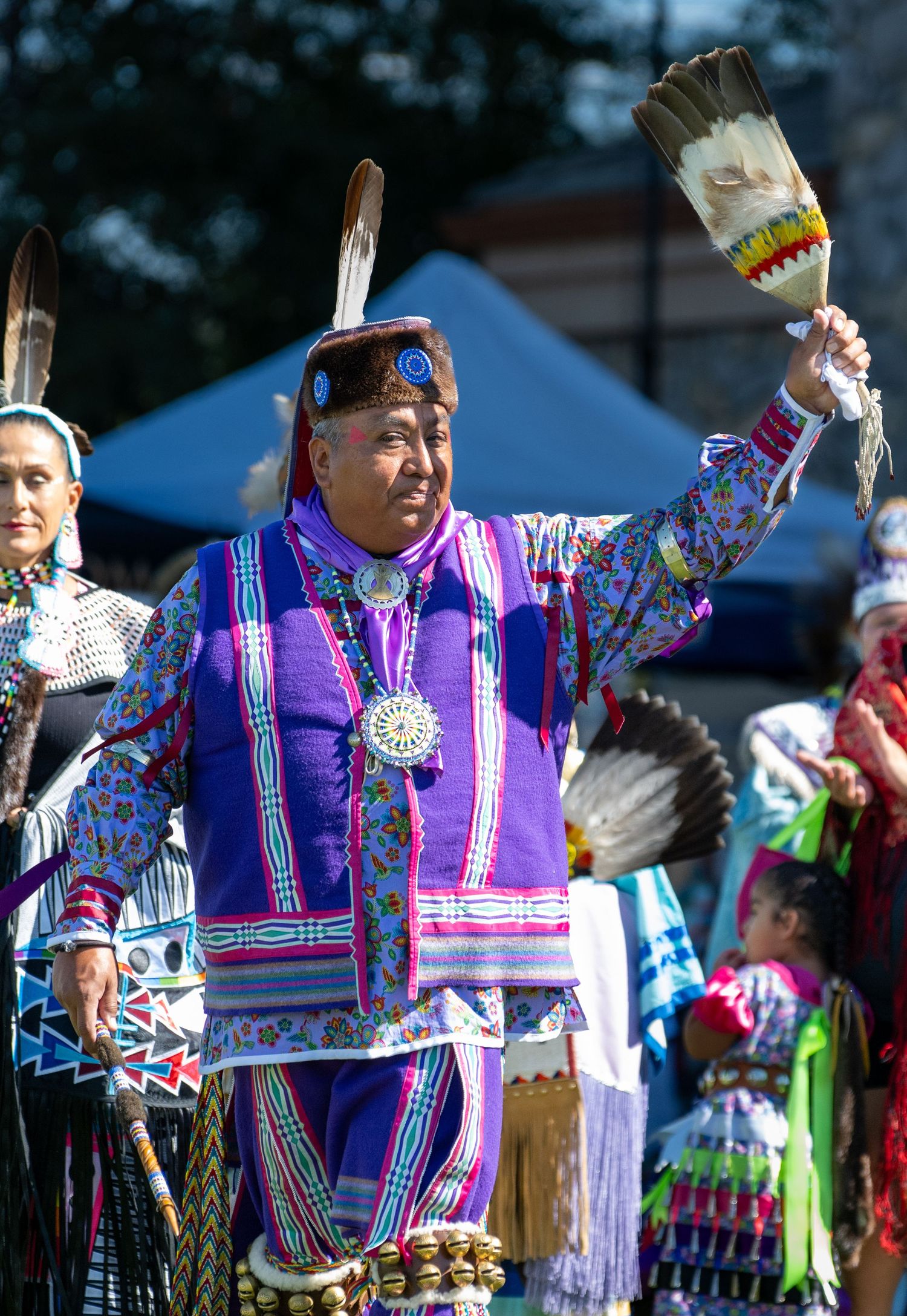 Dancers in vibrant purple and turquoise traditional regalia perform at an outdoor powwow event.
