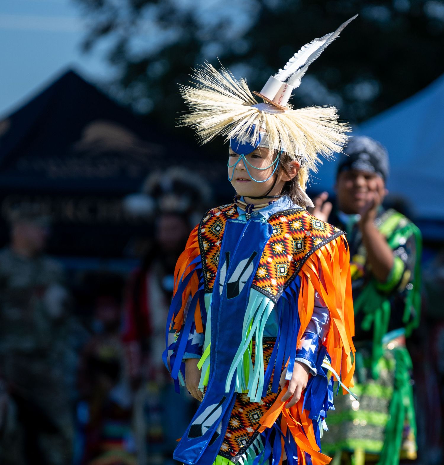 A Native American dancer in bright orange and blue regalia performs at a festival.
