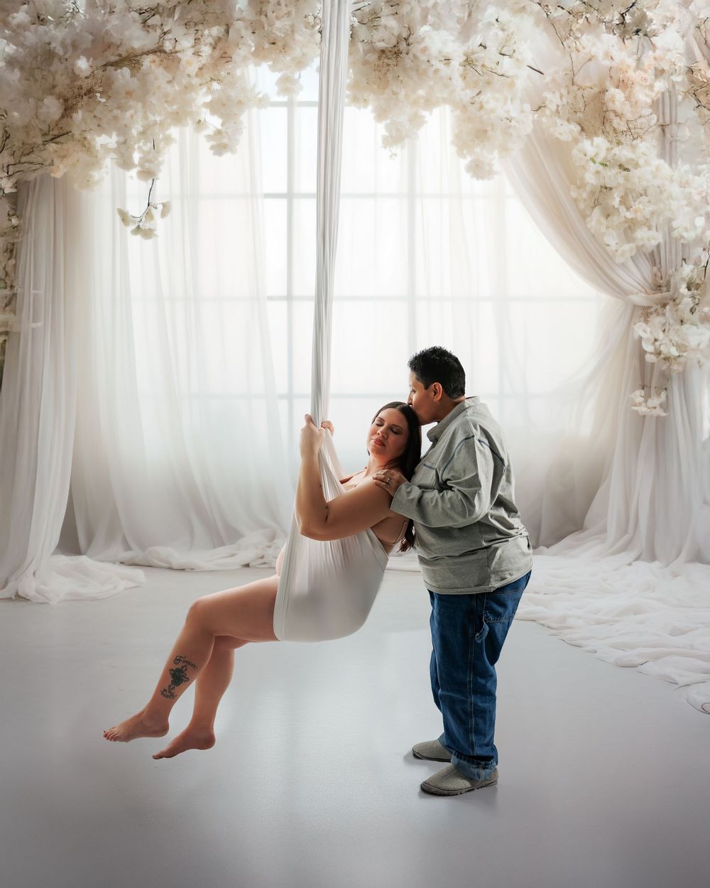 Romantic indoor portrait with white floral decorations and flowing curtains in a dreamy studio setting.