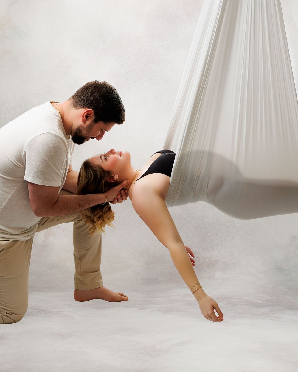 Two dancers perform an intimate contemporary dance move with aerial silk drapes in a minimalist white studio setting.