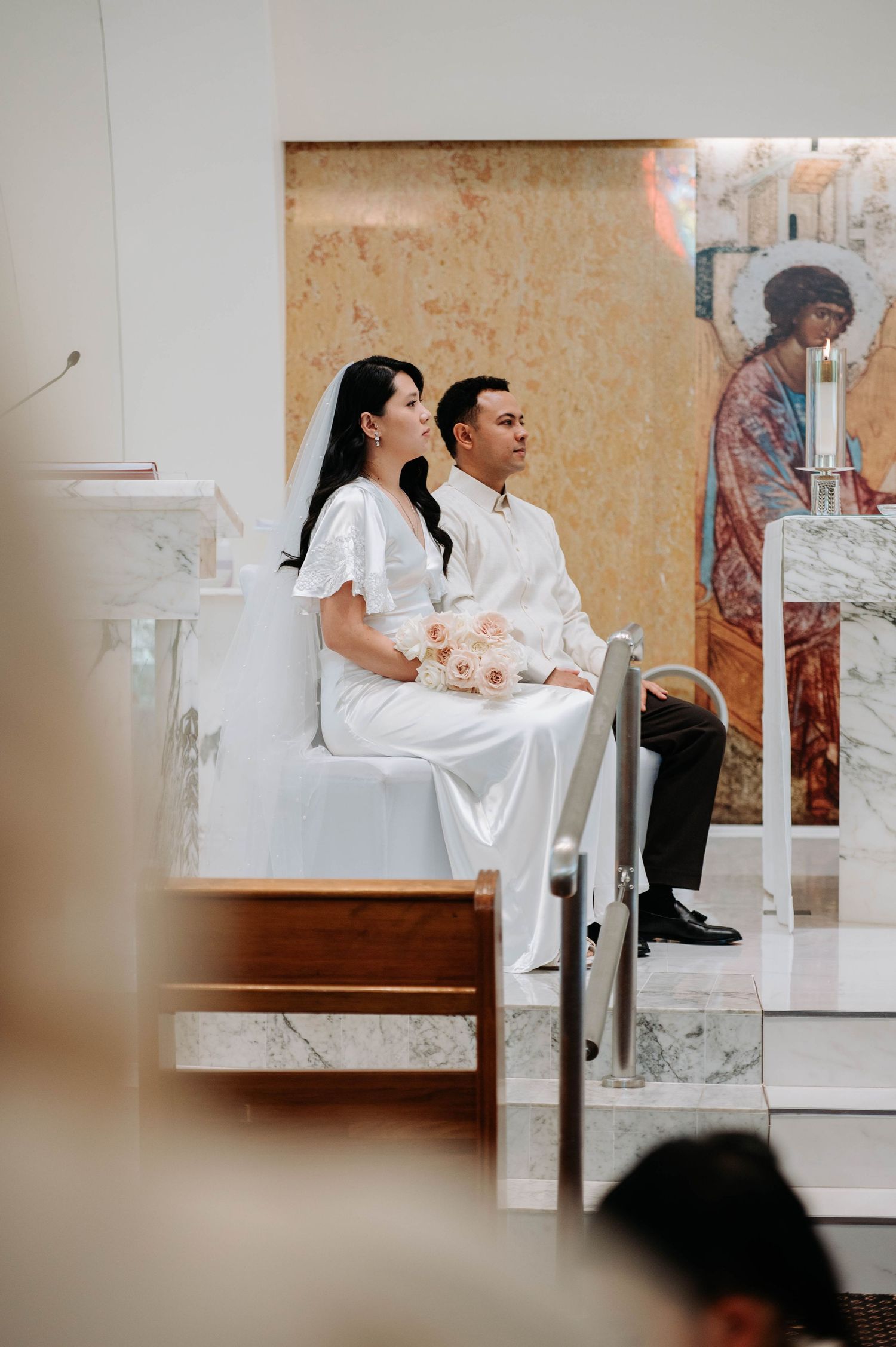 A couple in white attire sits together during a church ceremony with religious artwork visible in the background.
