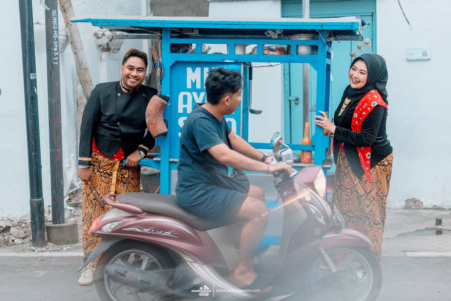 Three people chat and laugh near a bright turquoise doorway with a parked scooter on a city street.