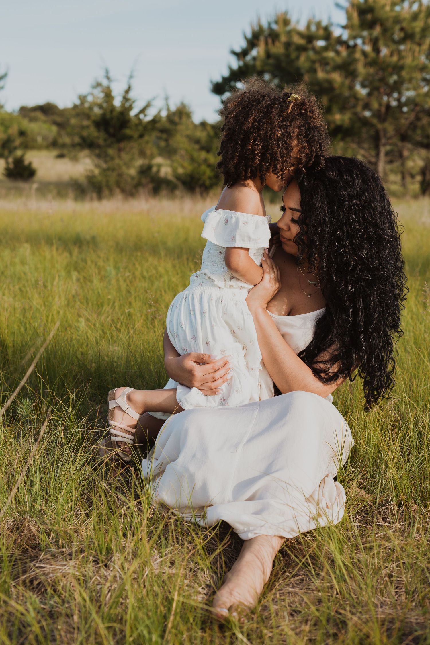 Mother and child in white dresses share a tender moment while sitting in a sunlit grassy meadow at golden hour.