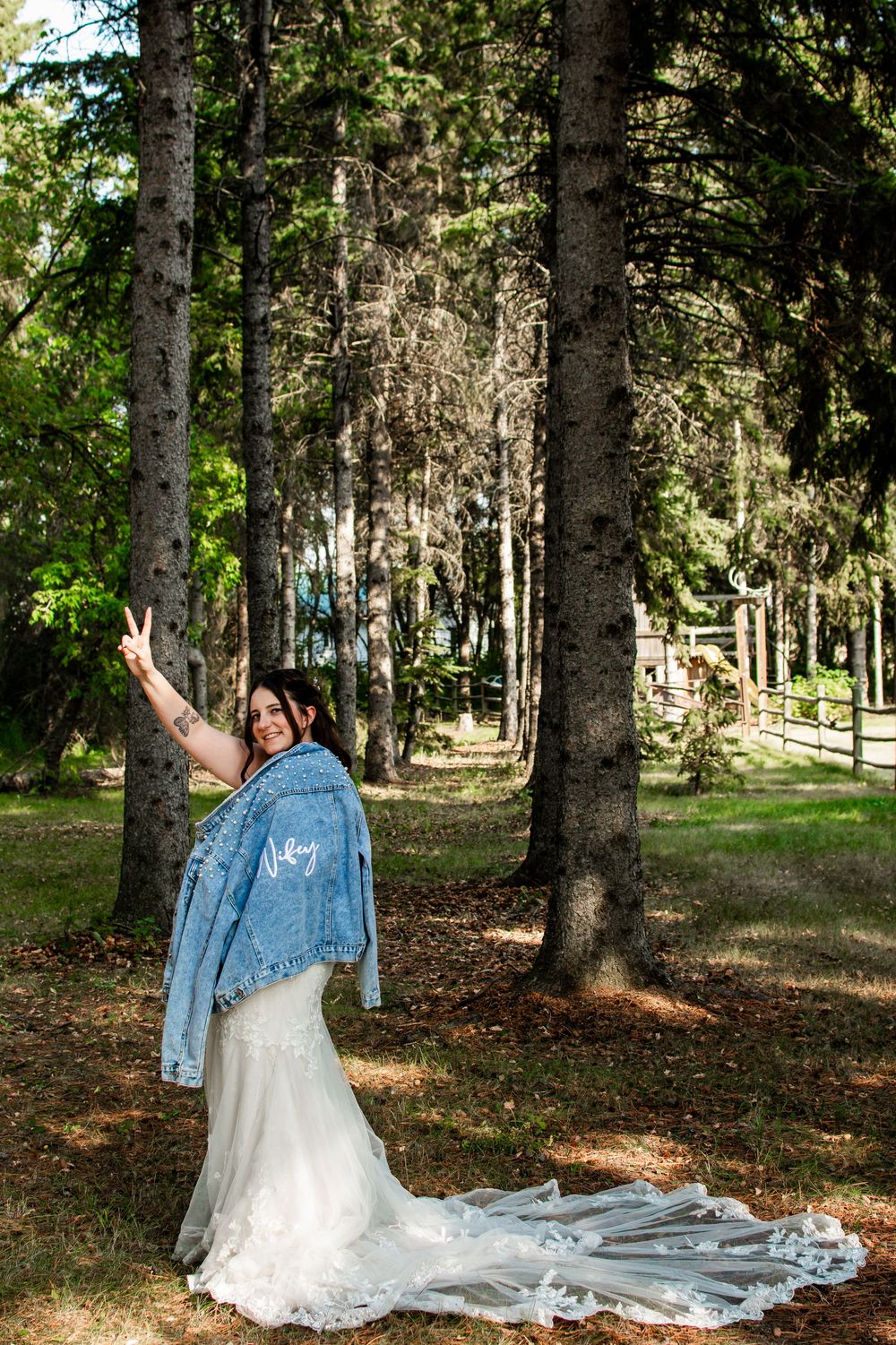 Person in wedding dress and denim jacket poses playfully in a wooded setting.