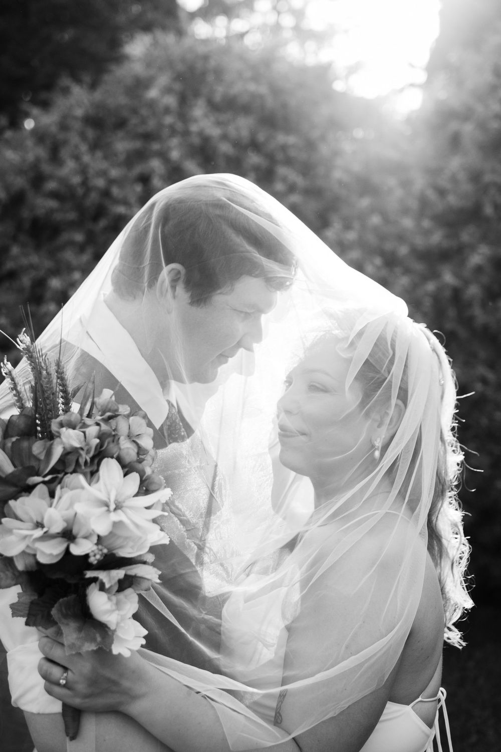 Romantic black and white portrait of newlyweds embracing under flowing veil with floral bouquet.