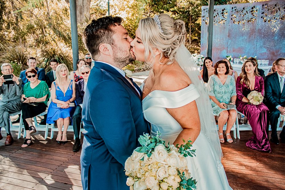 A romantic first kiss at an outdoor wedding ceremony with guests watching in colorful attire.
