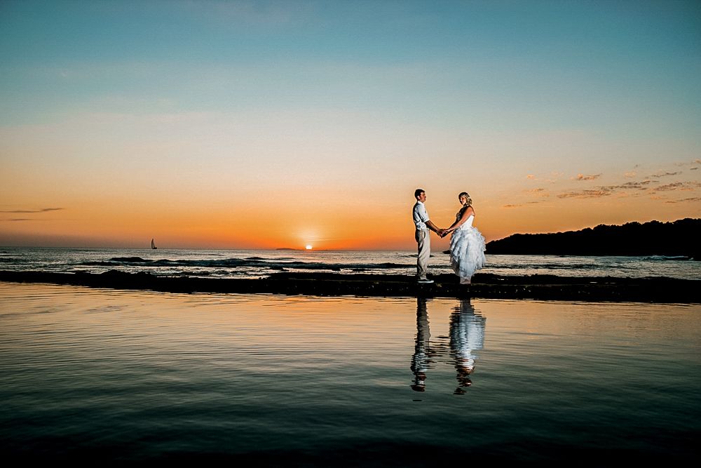 Silhouetted couple holding hands on beach at sunset with reflection in wet sand.