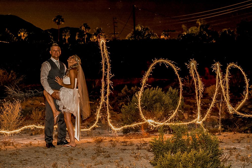 Couple stands beside illuminated LOVE sign made of sparklers during nighttime outdoor wedding photography.