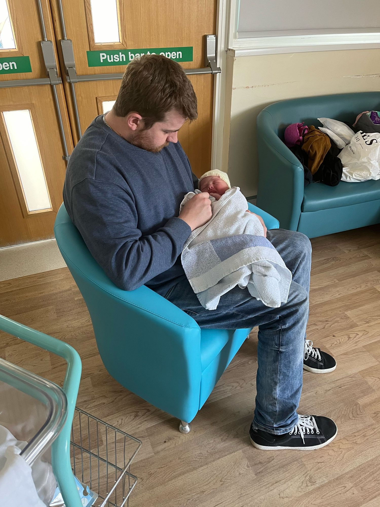 A person in a gray sweater sits in a turquoise armchair while holding an infant wrapped in white blankets.