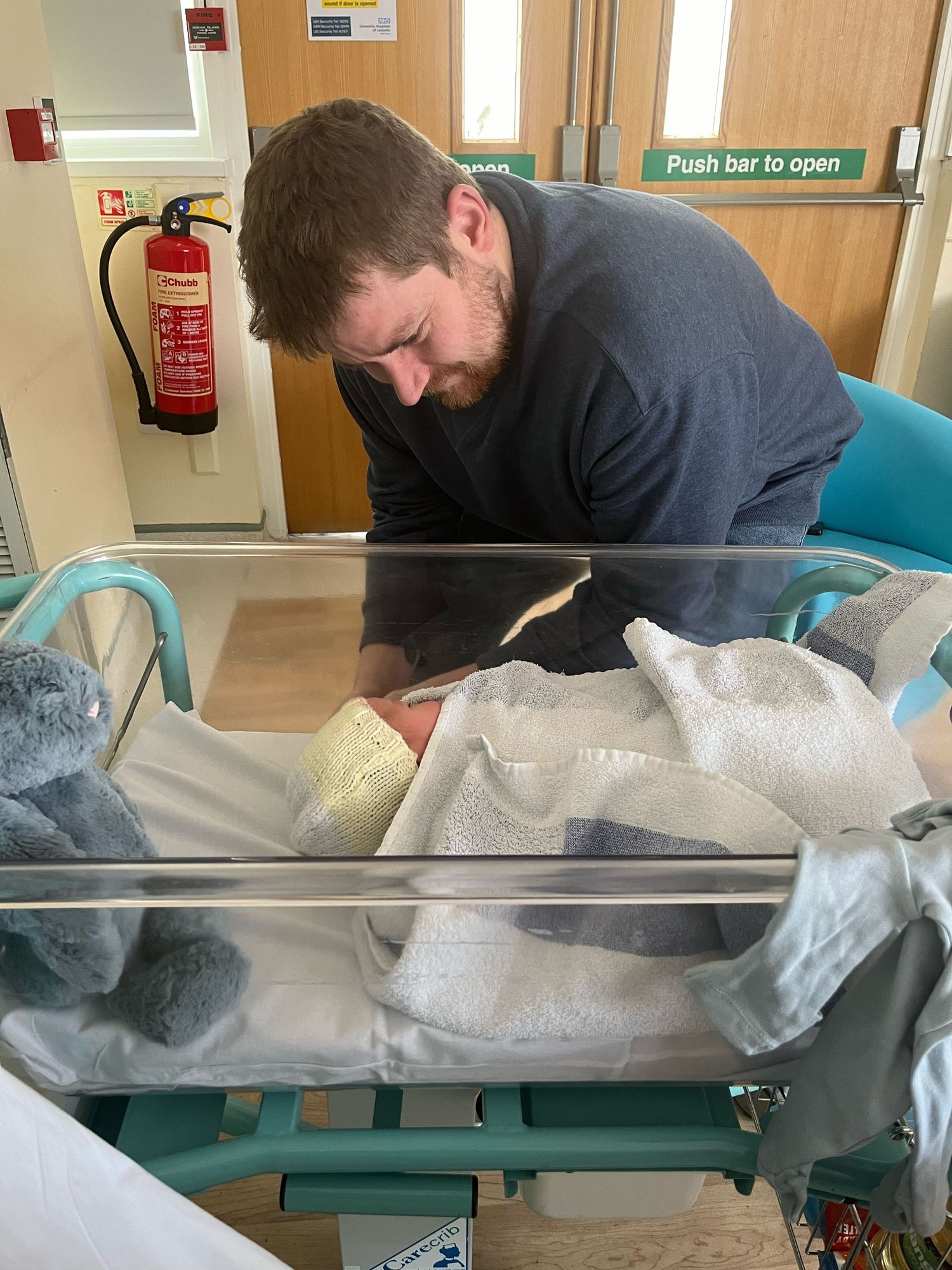 A person wearing a dark sweater leans over a hospital bassinet containing swaddled blankets and medical supplies.