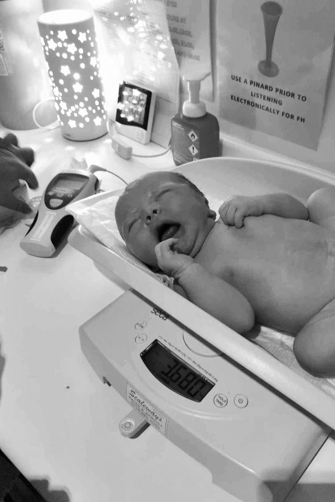 A newborn baby lies on a digital scale during their first medical checkup, with a glowing lamp visible in the background.