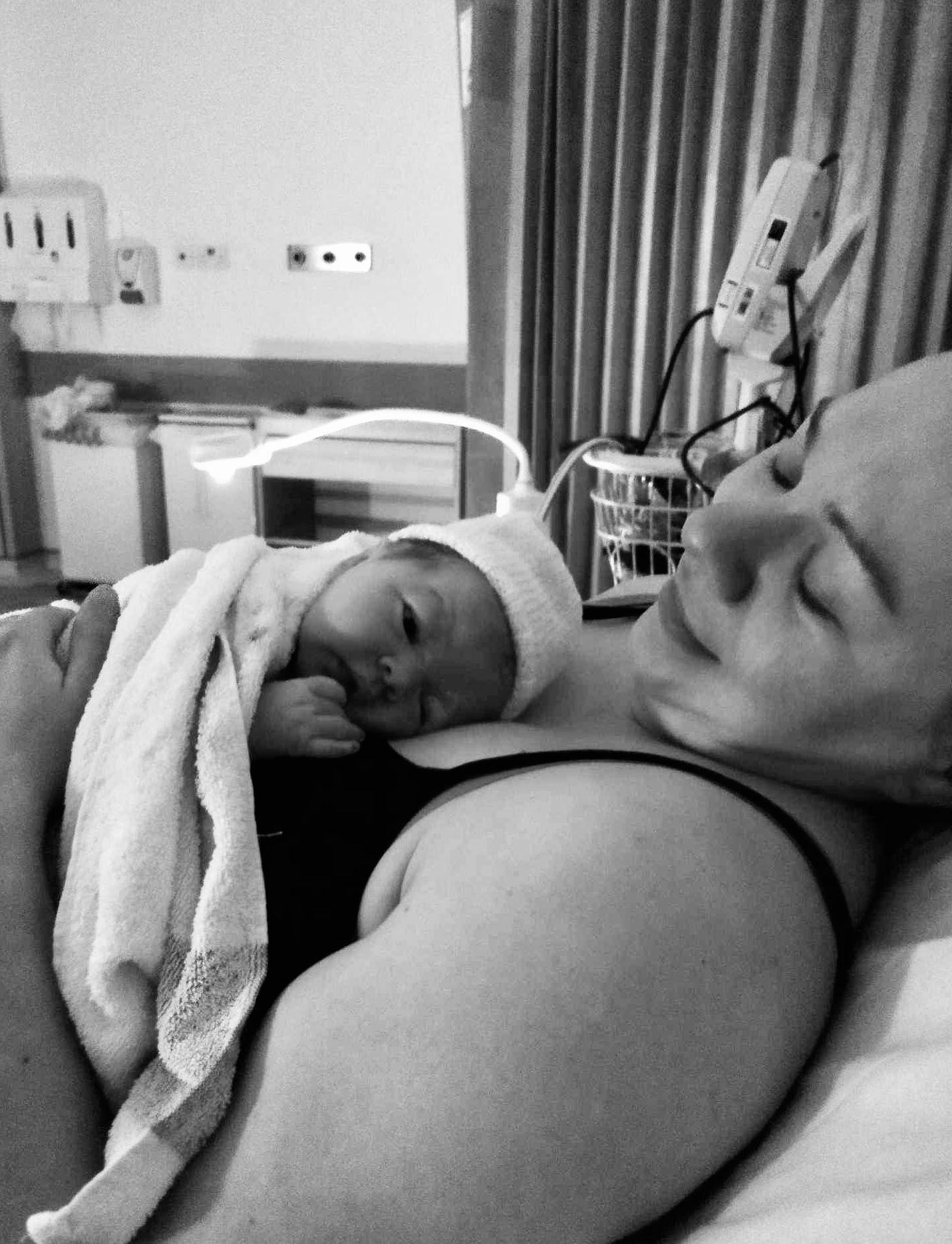 A newborn baby in white swaddle rests on an adult's chest in a black and white hospital scene.