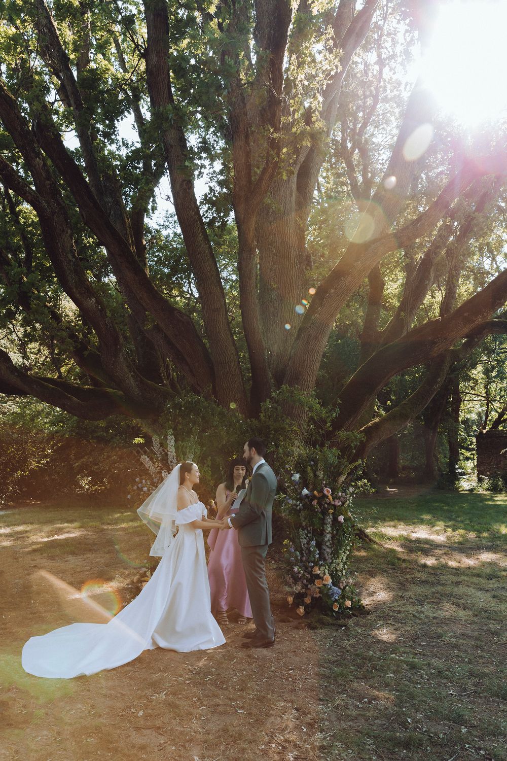 Sunlit wedding ceremony under large oak trees with streaming golden light.