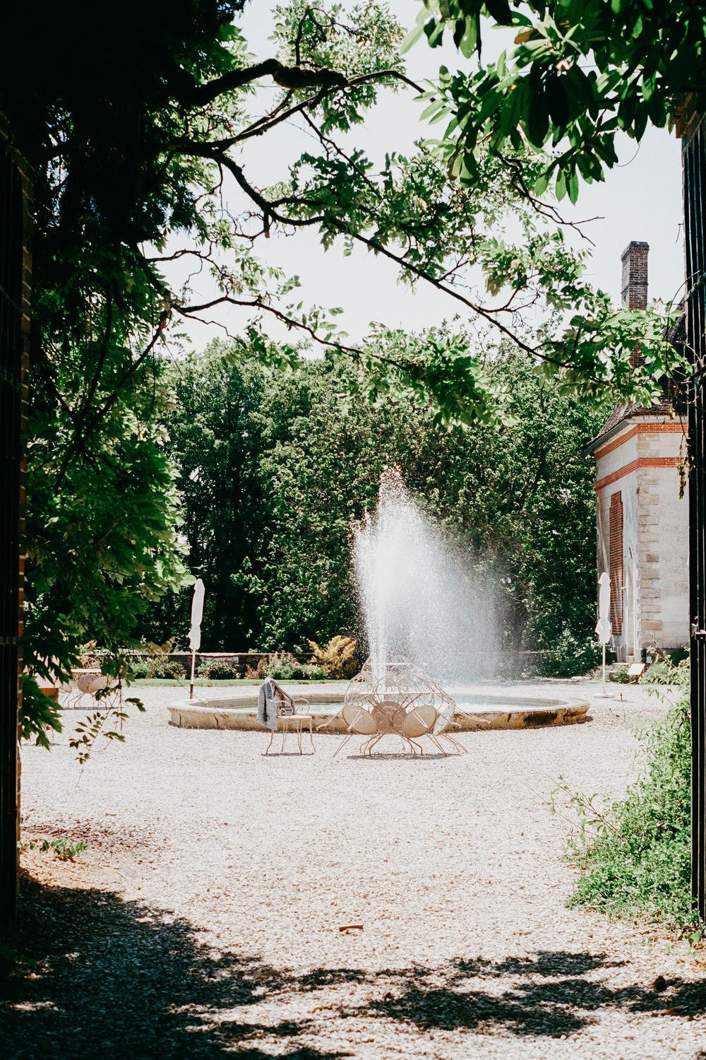 Garden fountain spraying water in a formal garden setting framed by green trees and architecture.